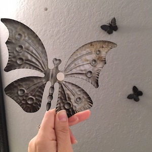 May include: A decorative metal butterfly with a distressed finish, held in front of a gray wall. The butterfly has detailed wing patterns and rivet-like accents. Two smaller butterfly wall decorations are also visible.