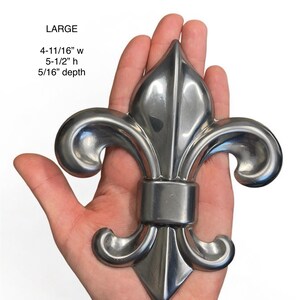 May include: A large, silver-colored fleur-de-lis, 4-11/16" wide, 5-1/2" high, and 5/16" deep. The decorative metal piece is held in a hand against a white background. The word "LARGE" is printed above the measurements.