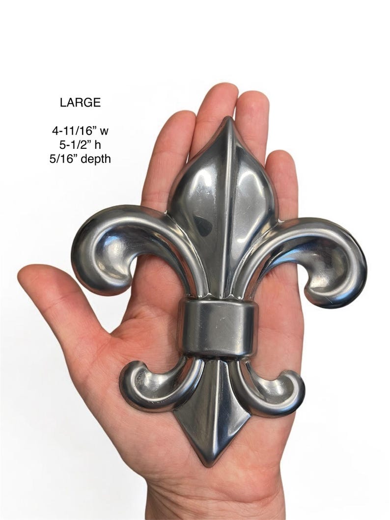 May include: A large, silver-colored fleur-de-lis, 4-11/16" wide, 5-1/2" high, and 5/16" deep. The decorative metal piece is held in a hand against a white background. The word "LARGE" is printed above the measurements.