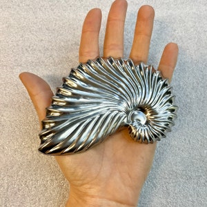 Puede incluir: Una escultura de concha de nautilus plateada pulida. La forma espiral de la concha está detallada con texturas acanaladas, que reflejan la luz. La escultura se sostiene en una mano, mostrando su tamaño y diseño. Un artículo decorativo.