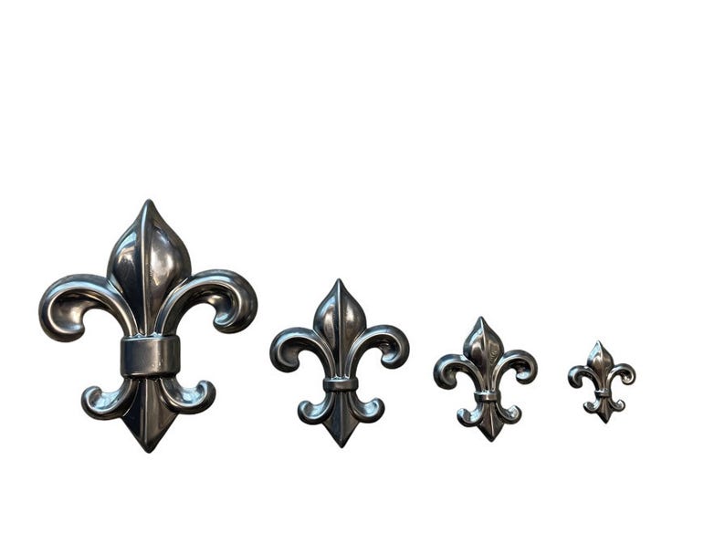 May include: Four silver-colored fleur-de-lis ornaments of varying sizes. The ornaments have a classic three-petal design with curved accents. The largest is on the left, with the smallest on the right.