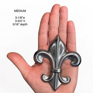 May include: A silver-colored fleur-de-lis, approximately 3-1/8" wide, 3-3/4" high, and 5/16" deep, is held in a hand. The word "MEDIUM" is above the measurements. The design features a stylized lily with three petals.