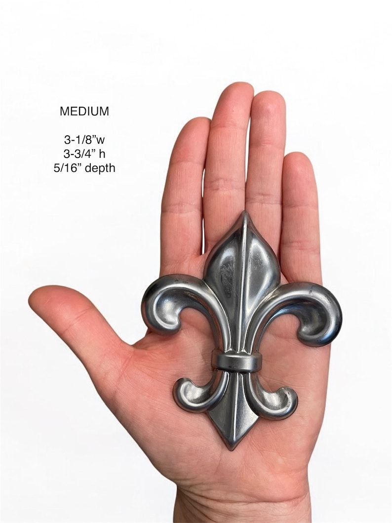 May include: A silver-colored fleur-de-lis, approximately 3-1/8" wide, 3-3/4" high, and 5/16" deep, is held in a hand. The word "MEDIUM" is above the measurements. The design features a stylized lily with three petals.