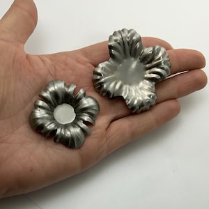 Puede incluir: Dos adornos de metal plateado en forma de flor. La flor más grande tiene cinco pétalos y la flor más pequeña tiene seis pétalos.