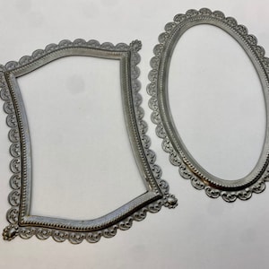 May include: Two silver metal picture frames with ornate scalloped edges. The frame on the left is a curved rectangle shape, and the frame on the right is an oval shape.