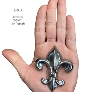 May include: A small, silver-colored fleur-de-lis ornament, 2-3/8" wide, 2-3/4" high, and 1/4" deep, is held in an open hand. The ornament has a detailed, symmetrical design with a pointed top and curved side elements. The word "SMALL" is above the measurements.