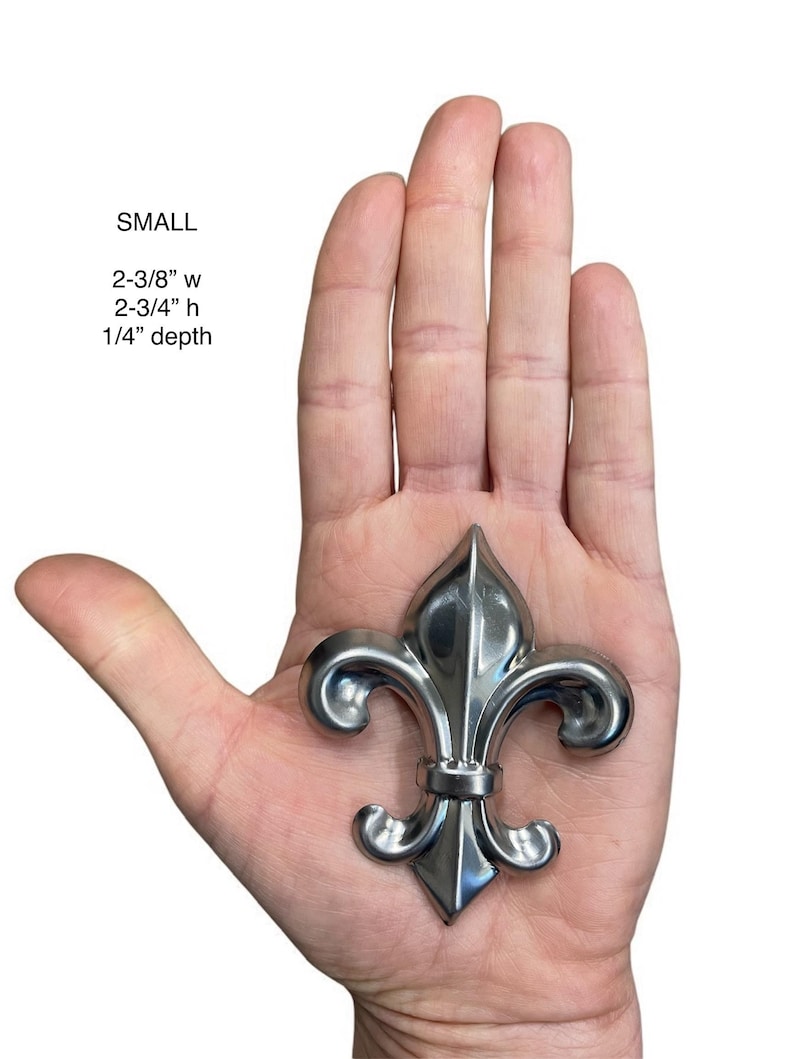 May include: A small, silver-colored fleur-de-lis ornament, 2-3/8" wide, 2-3/4" high, and 1/4" deep, is held in an open hand. The ornament has a detailed, symmetrical design with a pointed top and curved side elements. The word "SMALL" is above the measurements.