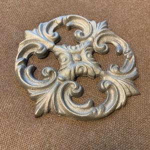 May include: A silver metal decorative accent with a swirling, ornate design. The accent has a four-leaf clover shape with a raised center.