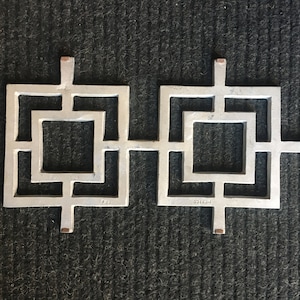 Puede incluir: Una decoración de pared de metal plateado con un diseño geométrico. El diseño presenta dos cuadrados con un cuadrado más pequeño dentro de cada uno. Los cuadrados están conectados por una barra horizontal. La decoración de pared está marcada con "MEXICO" en el lado derecho y "701" en el lado izquierdo.