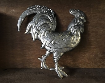 Può includere: Scultura di gallo color argento con dettagli di piume. Il gallo di metallo ha una superficie strutturata. Il gallo è su una superficie di legno. Oggetto decorativo.