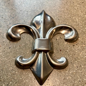 May include: A silver-colored fleur-de-lis emblem with a polished finish. The emblem features three stylized petals, a central vertical element, and curved side elements, all connected by a horizontal band.