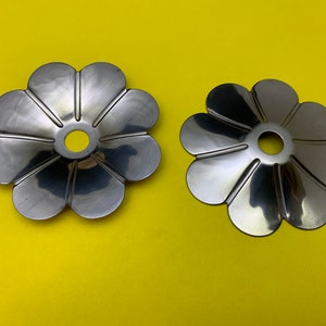 May include: Two silver metal flower-shaped decorative pieces with a hole in the centre. The pieces are on a yellow background.