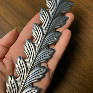 May include: A silver-colored, leaf-shaped decorative element. The metal leaf has detailed veining and a textured surface. The leaf is held in a hand, showcasing its size and design. The item is likely for home decor.