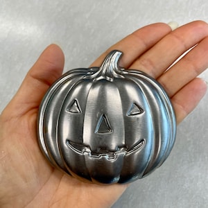 Peut inclure: Une citrouille en métal argenté avec un visage de citrouille sculpté. La citrouille est à peu près de la taille de la main d'une personne.