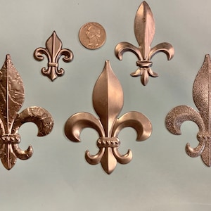 May include: Five bronze-colored fleur-de-lis ornaments of varying sizes. The fleur-de-lis are arranged on a light green surface, with a U.S. quarter coin for size comparison. The ornaments have detailed textures and a metallic finish.