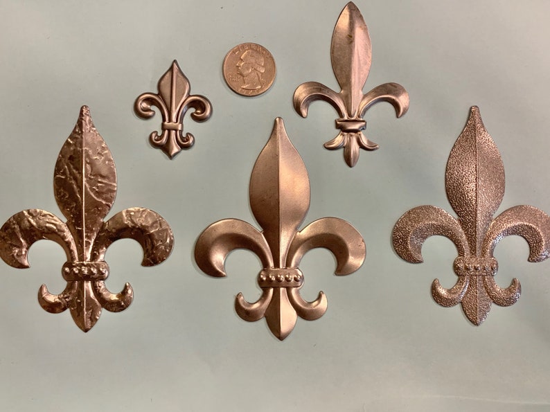 May include: Five bronze-colored fleur-de-lis ornaments of varying sizes. The fleur-de-lis are arranged on a light green surface, with a U.S. quarter coin for size comparison. The ornaments have detailed textures and a metallic finish.