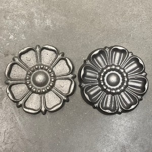 Op de afbeelding: Twee zwarte metalen decoratieve stukken in de vorm van een bloem met een verhoogde middelste cirkel en een getextureerd oppervlak.