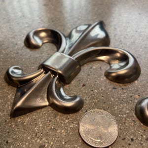 May include: A silver-colored fleur-de-lis emblem with a quarter coin for size comparison. The emblem has a detailed, metallic finish and a classic design. The coin shows the profile of George Washington.