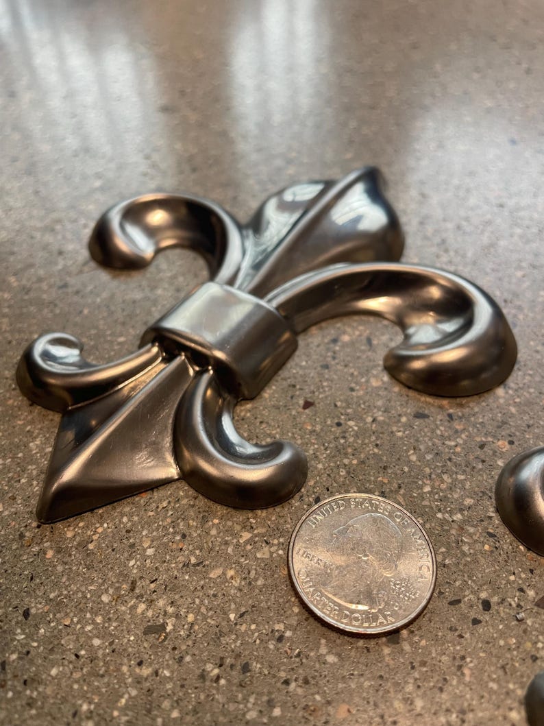 May include: A silver-colored fleur-de-lis emblem with a quarter coin for size comparison. The emblem has a detailed, metallic finish and a classic design. The coin shows the profile of George Washington.