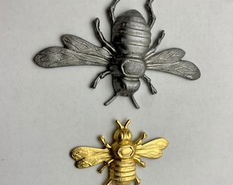 May include: Two metal bee figurines, one silver and one gold. The silver bee is larger and has more detail. The gold bee is smaller and has a more simple design.