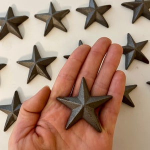 May include: A collection of ten cast iron five-pointed stars in a dark gray color. The stars are scattered on a white surface with one star being held in a hand.