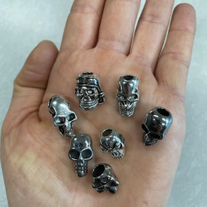 May include: Seven silver skull-shaped beads with different designs. The beads have a hole through the top for stringing.