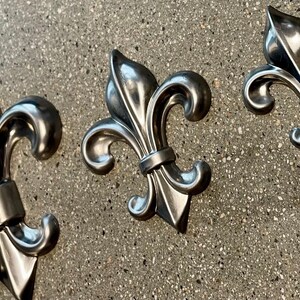 May include: Close-up of three silver fleur-de-lis ornaments. The ornaments are made of a shiny metal and are arranged diagonally across the frame. The background is a speckled gray surface.