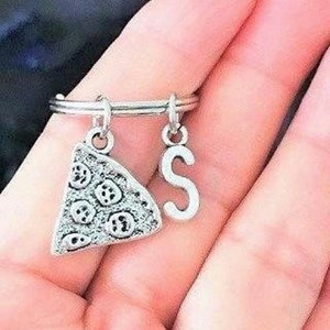 May include: A silver-toned keychain featuring a pizza slice charm and the letter "S" charm. The pizza slice has a detailed design with small circles representing toppings. The keychain is held in a person's hand.