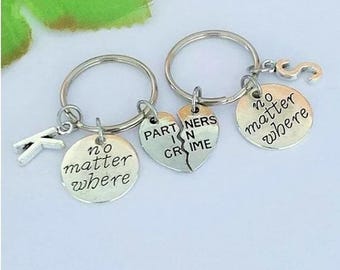 Puede incluir: Dos llaveros plateados con colgantes redondos y en forma de corazón. Un llavero tiene el texto "no matter where" y un dije con la letra "K". El otro llavero tiene un colgante de corazón roto con el texto "PARTNERS IN CRIME".