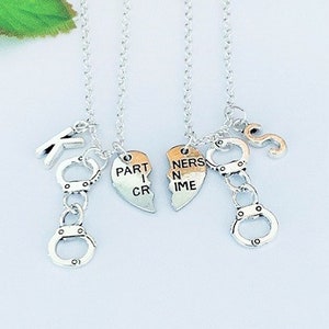 May include: Two silver-toned necklaces with matching pendants. Each necklace features a small handcuff charm, a letter charm, and a heart-shaped pendant that reads "PARTNER IN CRIME" when joined. The chains are delicate and the background is light.