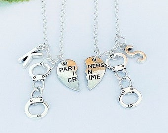 May include: Two silver-toned necklaces with matching pendants. Each necklace features a small handcuff charm, a letter charm, and a heart-shaped pendant that reads "PARTNER IN CRIME" when joined. The chains are delicate and the background is light.
