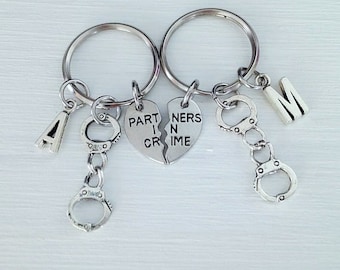 May include: Two silver keychains with matching broken heart charms that say "Partners in Crime" and "Part in Crime". Each keychain also has a silver letter charm, a silver handcuff charm, and a silver key ring.