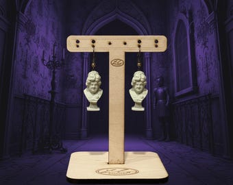 May include: A pair of white bust earrings with a vintage aesthetic, displayed on a wooden earring stand. The earrings depict a classical bust design. The stand is made of light-colored wood and has a rectangular base.