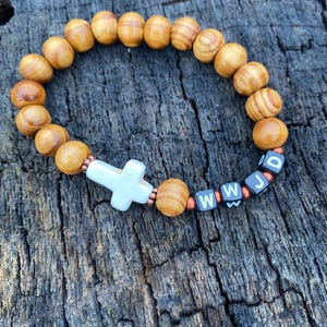 May include: A wooden bead bracelet with a white cross and the letters "WWJD" on black and white beads. The bracelet features light brown wooden beads and is displayed on a weathered wooden surface.
