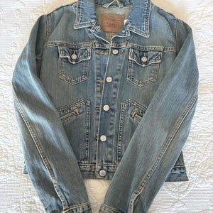 May include: A light-wash denim jacket with a classic design. Features include a collared neckline, button-down front, chest pockets, and buttoned cuffs. The jacket has a faded appearance and a brand label on the back of the collar.