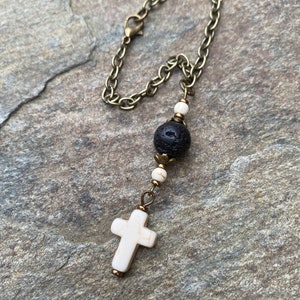 May include: A decorative hanging ornament featuring a bronze-toned metal chain attached to a series of beads. The beads include two small white marbled spheres, a larger porous black lava stone bead, and a white cross-shaped pendant at the base.