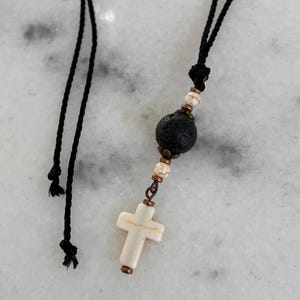 May include: A necklace featuring a black cord with a central pendant consisting of a porous black lava bead, two small white and brown marbled beads, and a white cross-shaped bead, all accented with small metallic copper-toned spacers.