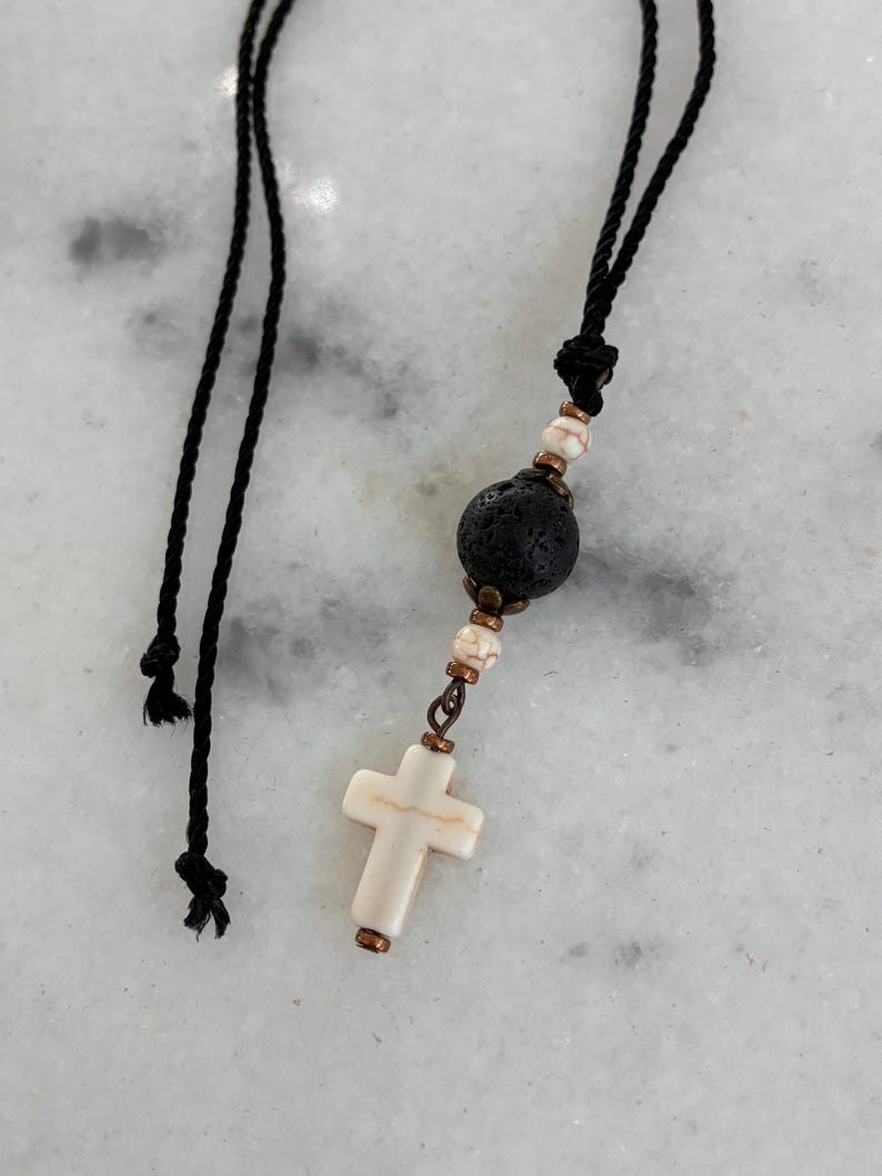 May include: A necklace featuring a black cord with a central pendant consisting of a porous black lava bead, two small white and brown marbled beads, and a white cross-shaped bead, all accented with small metallic copper-toned spacers.