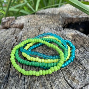 Peut inclure: Un ensemble de cinq bracelets en perles de différentes nuances de vert et de bleu. Les bracelets sont empilés les uns sur les autres, créant un effet dégradé.