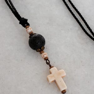 May include: A necklace featuring a black cord, a large porous black lava bead, two smaller white and brown marbled beads, and a white marbled cross pendant. The beads are accented with small bronze-toned metal spacers and connectors.