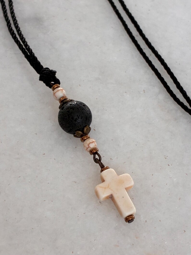 May include: A necklace featuring a black cord, a large porous black lava bead, two smaller white and brown marbled beads, and a white marbled cross pendant. The beads are accented with small bronze-toned metal spacers and connectors.