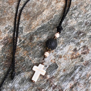 May include: A necklace featuring a black cord, a white cross pendant with brown veining, a large porous black lava bead, and two smaller white beads with brown veining, all accented with small metallic bronze-toned spacers and findings.