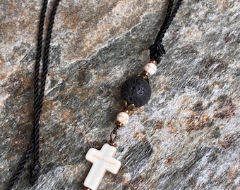May include: A necklace featuring a black cord, a white cross pendant with brown veining, a large porous black lava bead, and two smaller white beads with brown veining, all accented with small metallic bronze-toned spacers and findings.