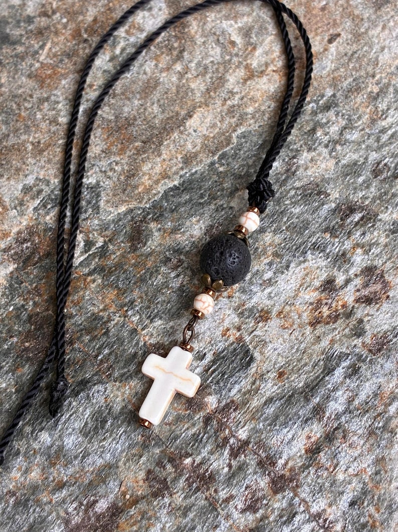 May include: A necklace featuring a black cord, a white cross pendant with brown veining, a large porous black lava bead, and two smaller white beads with brown veining, all accented with small metallic bronze-toned spacers and findings.