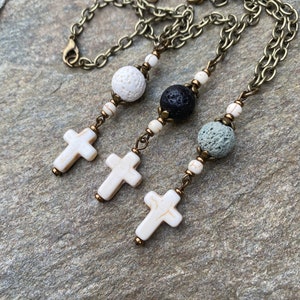 May include: Three necklaces featuring antique bronze chains, each with a cross pendant made of white stone with brown veining. The designs include porous lava beads in white, black, and grey, accented by small white stone beads and bronze metal spacers.