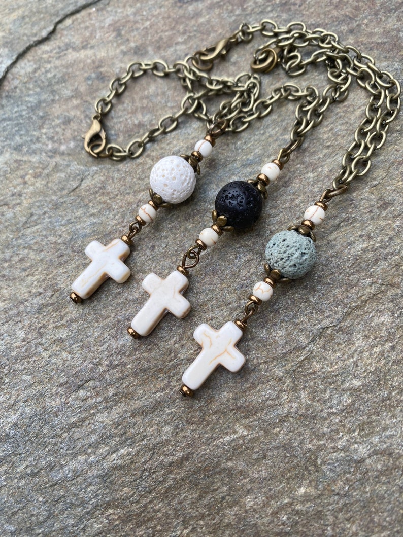 May include: Three necklaces featuring antique bronze chains, each with a cross pendant made of white stone with brown veining. The designs include porous lava beads in white, black, and grey, accented by small white stone beads and bronze metal spacers.