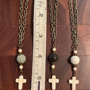 May include: Three bronze-toned chain charms featuring a white marble-style cross, two small white marble-style beads, and one larger porous bead in gray, black, or white. A white measuring tape shows the charms are approximately 5.5 inches long.