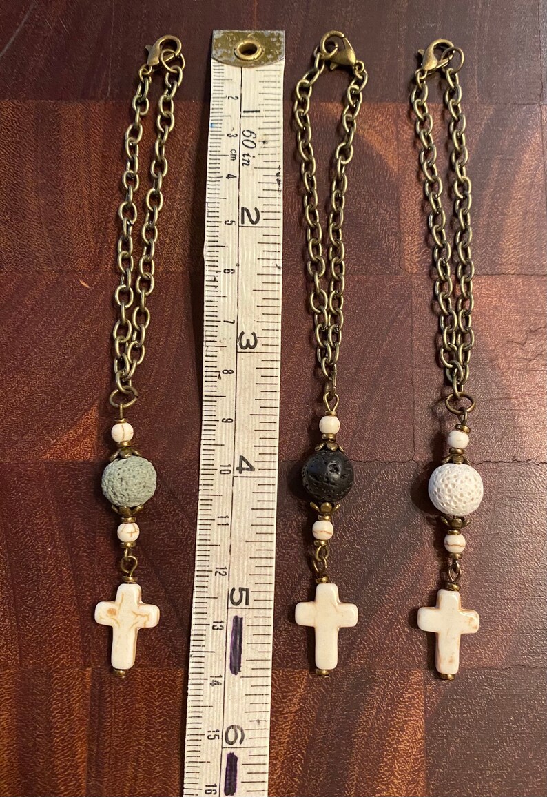May include: Three bronze-toned chain charms featuring a white marble-style cross, two small white marble-style beads, and one larger porous bead in gray, black, or white. A white measuring tape shows the charms are approximately 5.5 inches long.