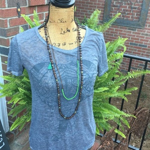 May include: A gray t-shirt with an elephant graphic print. The shirt is layered with two necklaces, one brown and one green.
