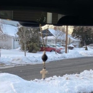 May include: A hanging rosary ornament featuring a gold-toned metal chain, a single large black spherical bead, and a small off-white cross pendant. The religious item is suspended from a vehicle rearview mirror, displaying a simple design.